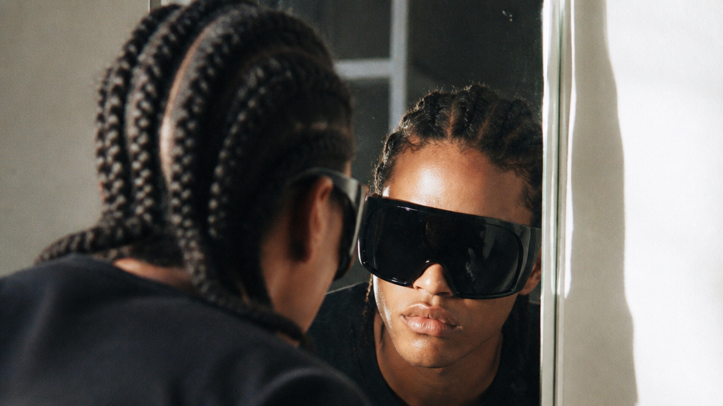 A person with braided hair looks into a mirror while wearing oversized black shield-style sunglasses or visor glasses. The reflection shows the front view of the black eyewear, and part of a bathroom sink and tiled wall are visible.